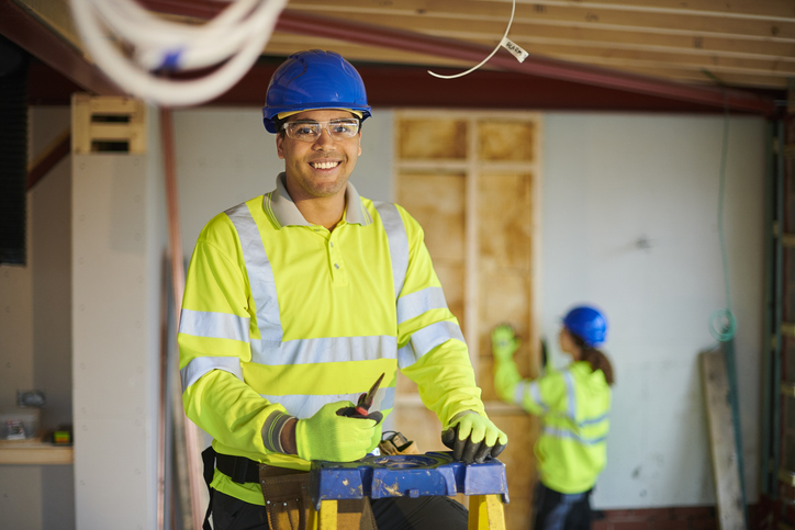Construction worker wearing safety equipment stands on a ladder at an indoor worksite.