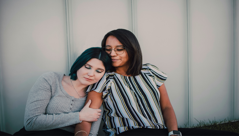 Two women sit together closely, one resting her head on the other’s shoulder in a tender moment