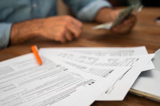 Close-up of tax documents and forms with a pen on a desk, while a person in the background counts cash