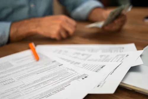 Close-up of tax documents and forms with a pen on a desk, while a person in the background counts cash