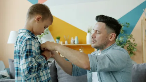 A father helps his young son button his shirt at home before heading out.