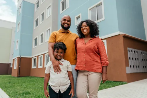 A smiling father, mother, and young son stand together outside a newly built multi-story residential apartment building with blue and brick exterior, representing a family benefiting from new affordable housing.
